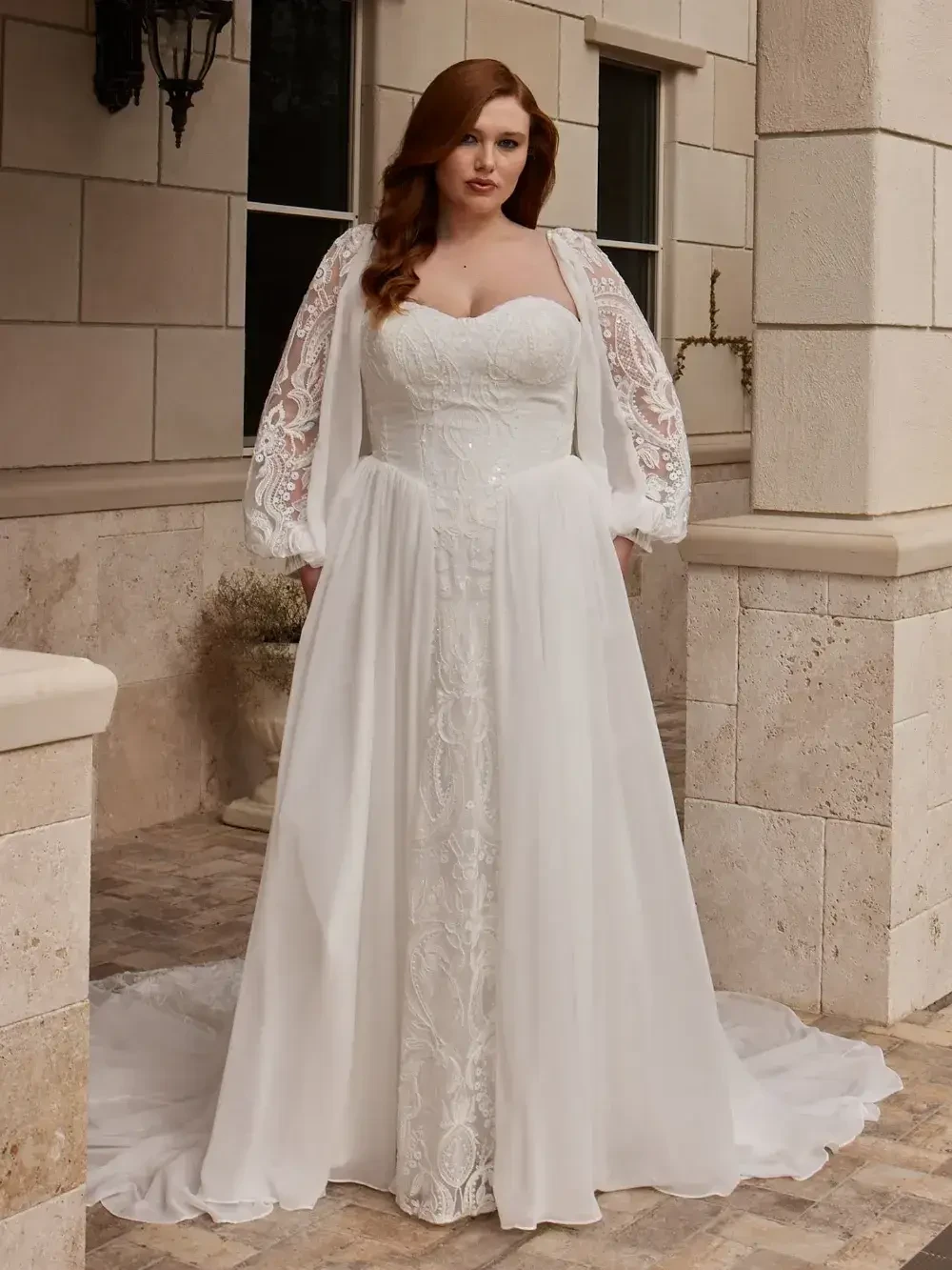 A woman stands confidently in a flowing white wedding dress with intricate lace and sheer long sleeves. She is outside on a stone patio, exuding elegance.