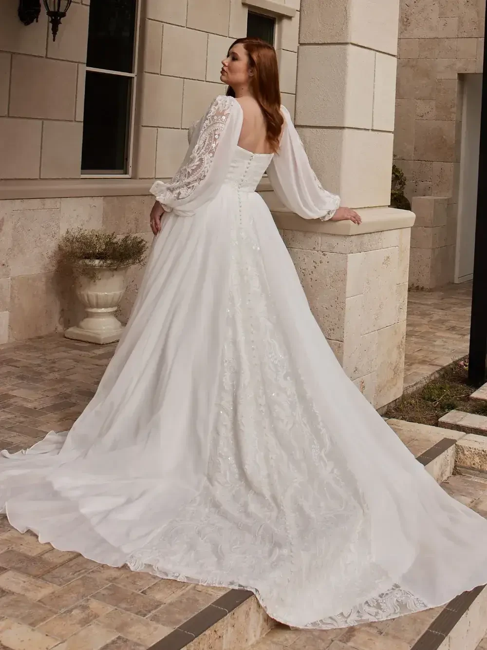 Bride in flowing white gown with lace sleeves and long train stands on a stone patio. The scene conveys elegance and tranquility against a stone building backdrop.