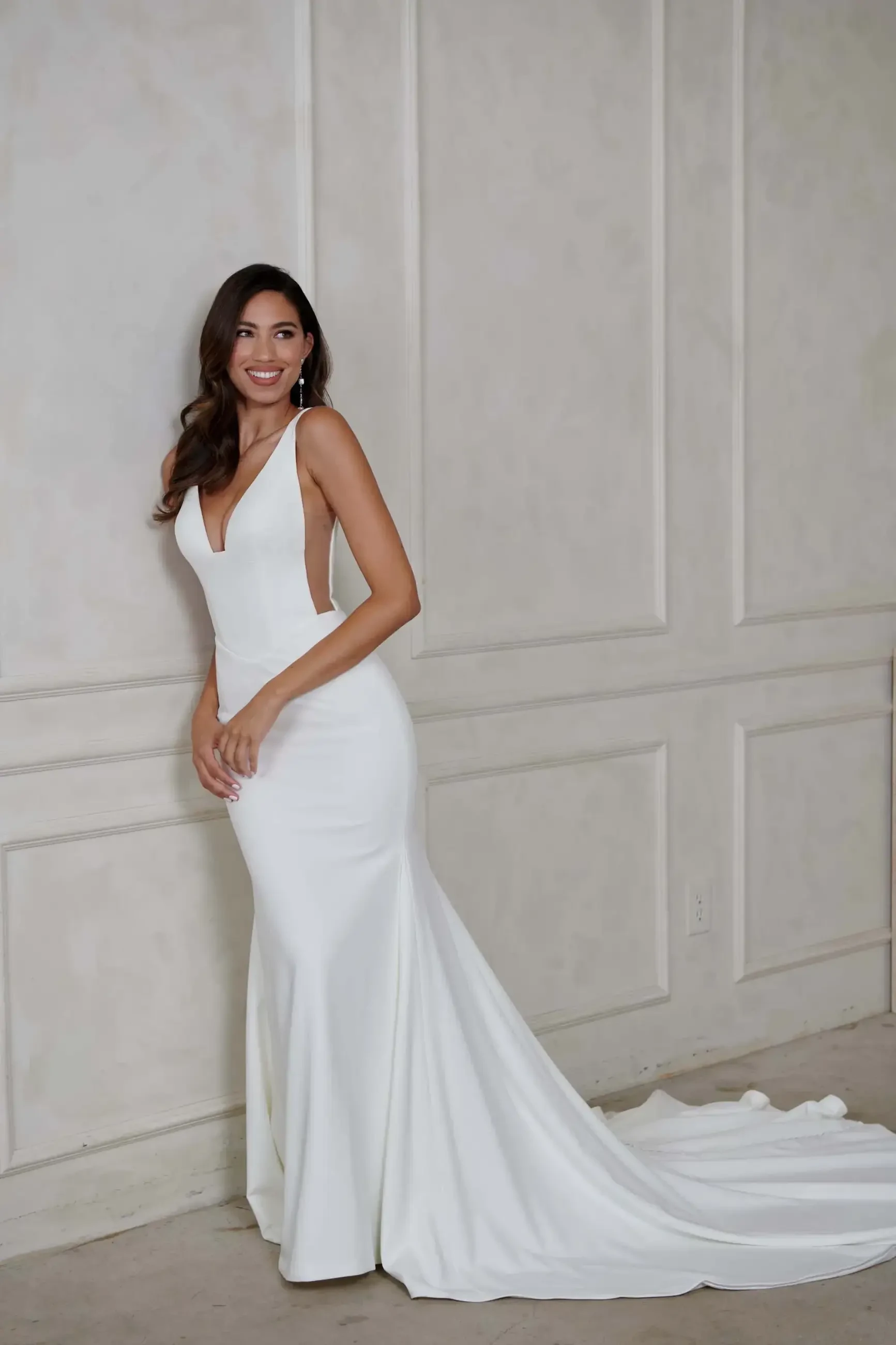 A woman in a sleek, elegant white gown leans against a beige paneled wall. She smiles warmly, conveying a sense of joy and elegance.