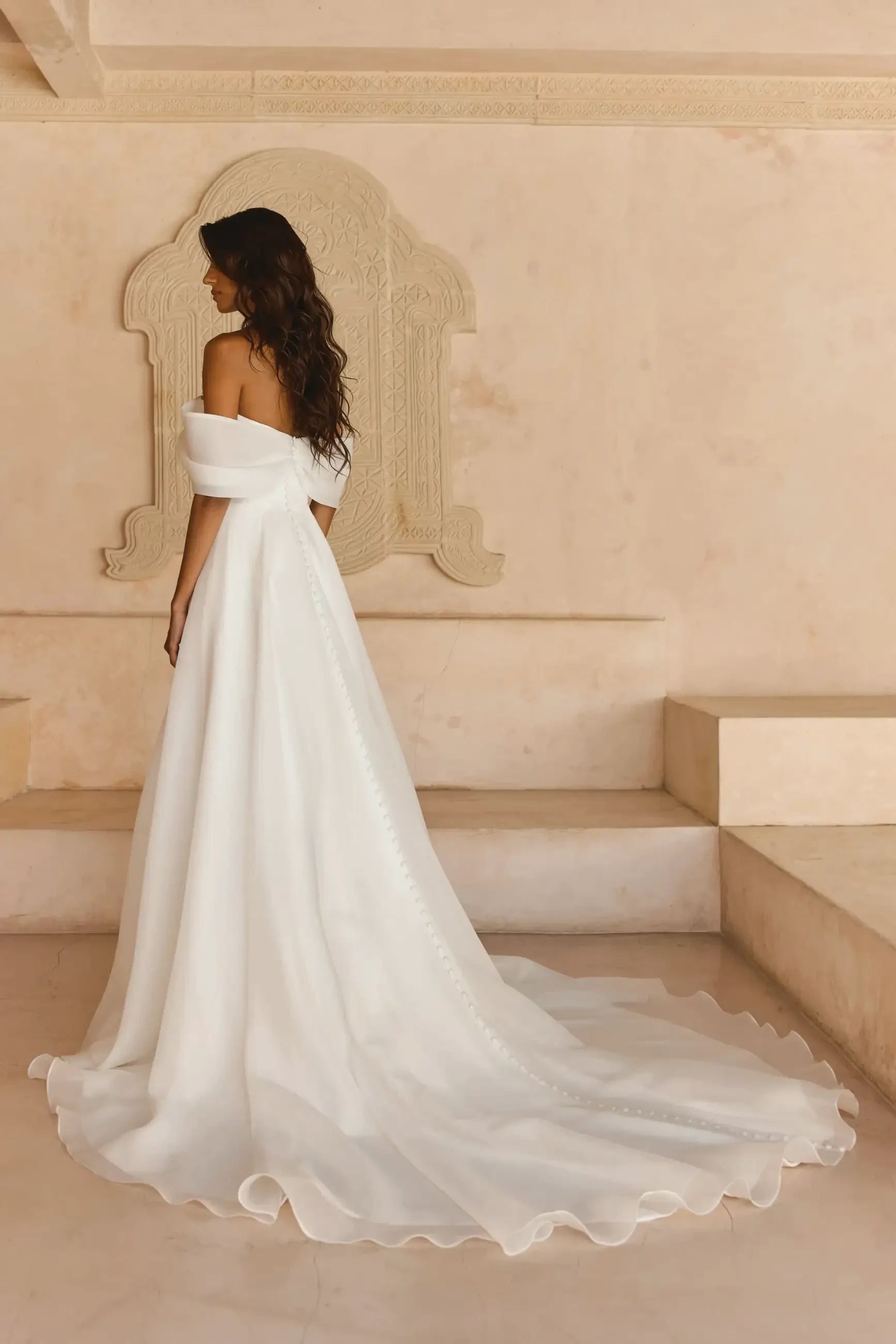 A woman in an elegant off-the-shoulder white wedding gown stands in a beige room with ornate wall details, creating a serene and graceful atmosphere.