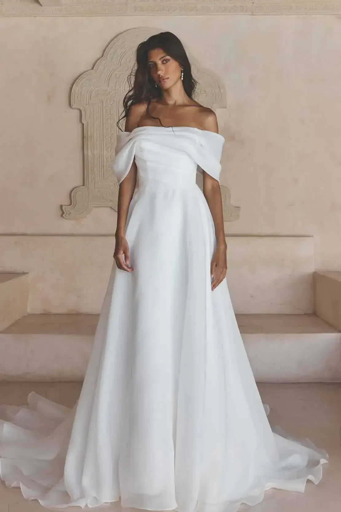 Woman in elegant off-shoulder white wedding dress stands gracefully in a neutral-toned room. The dress flows to the floor, exuding sophistication and calm.