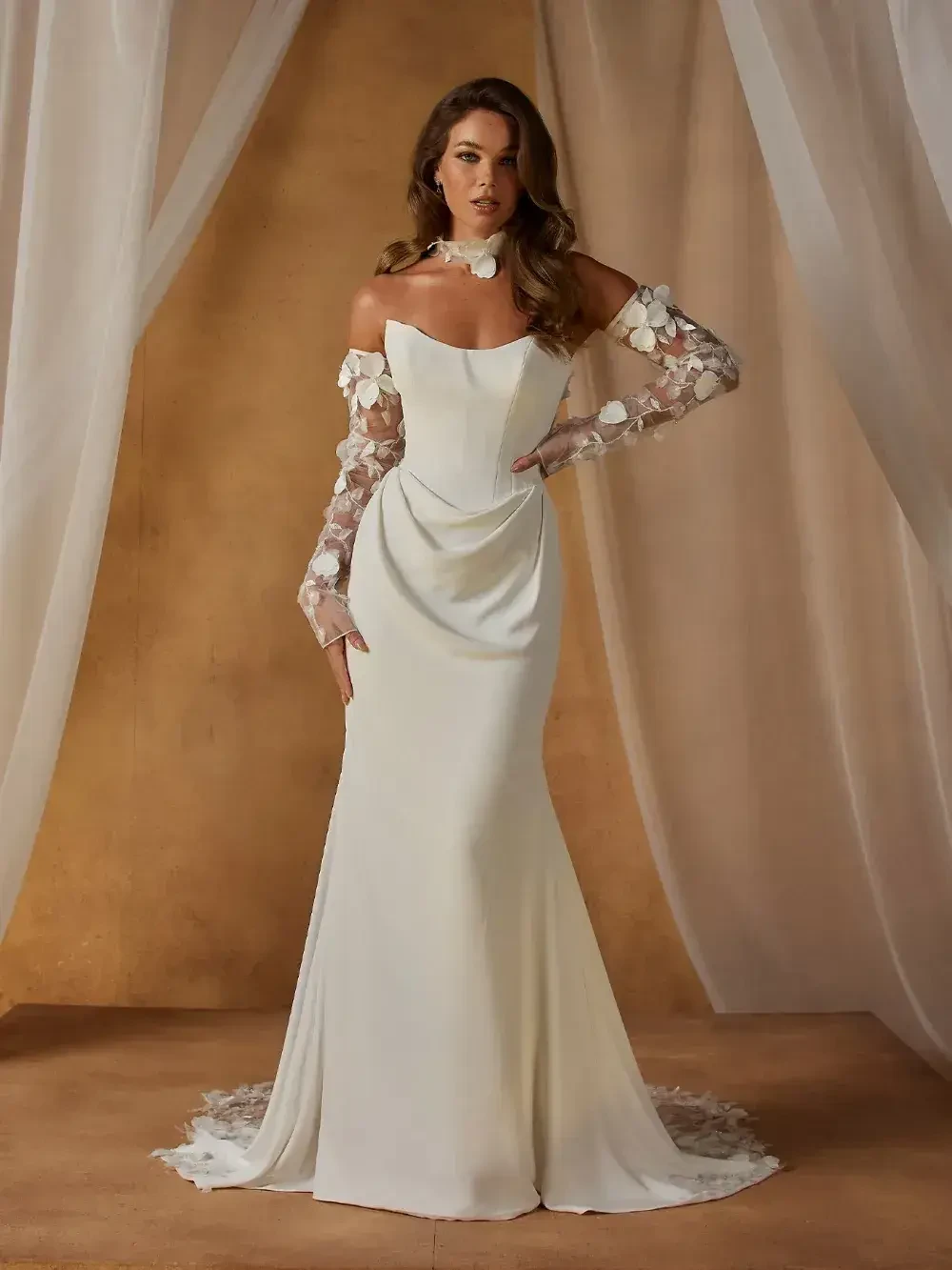Elegant woman in a flowing off-shoulder white gown with sheer floral sleeves poses against tan backdrop with soft lighting, conveying grace and sophistication.