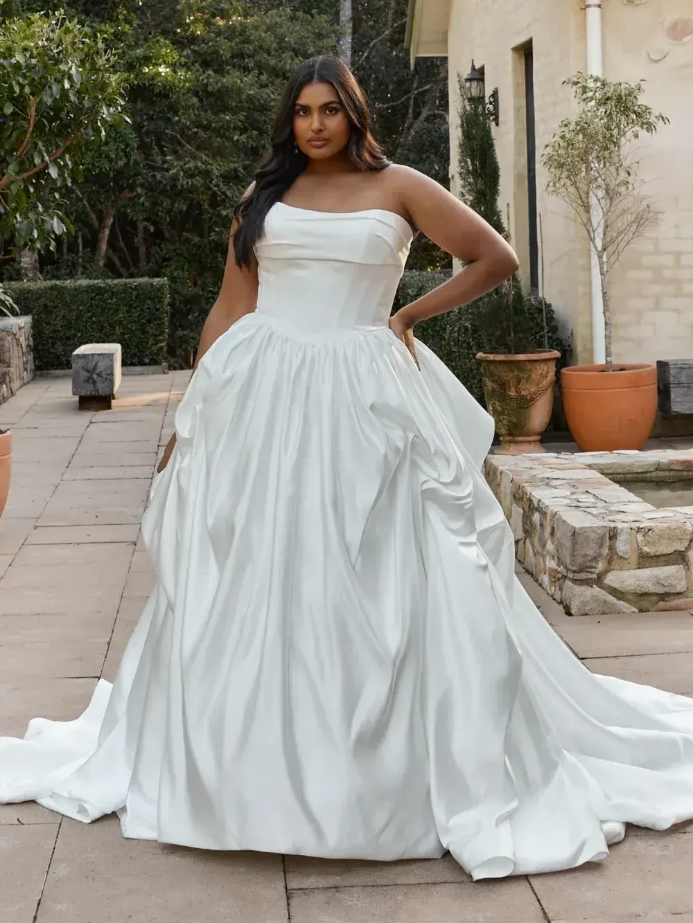 A woman in a strapless white gown standing outdoors, with greenery and stone features in the background.