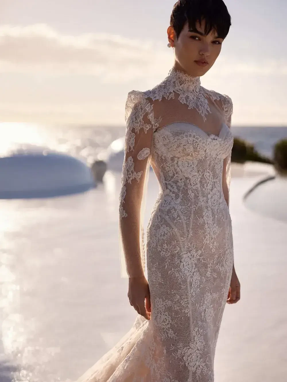 A woman in an elegant lace wedding gown poses outdoors by the shimmering sea. The setting sun casts a warm glow, evoking romance and serenity.