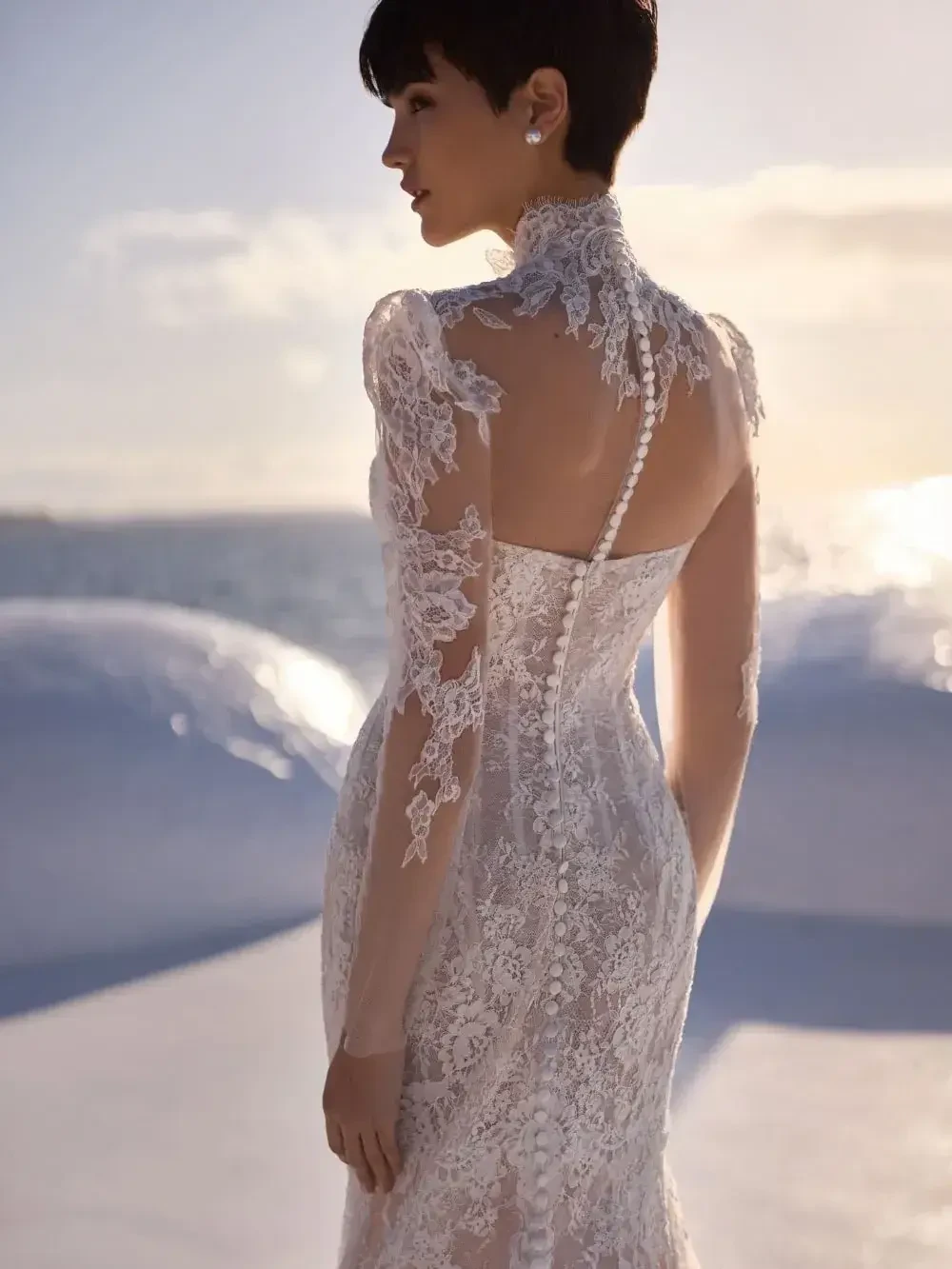 A woman stands by the ocean at sunset, wearing an elegant lace bridal gown with intricate details and a high collar, exuding a serene and romantic vibe.