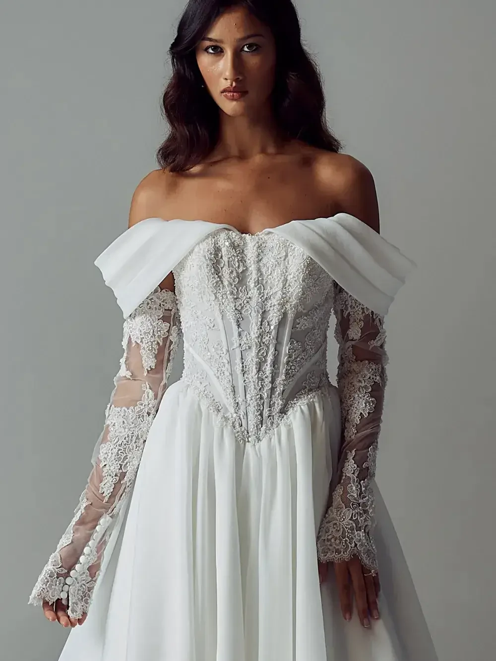 Woman in an elegant, off-shoulder wedding dress with delicate lace on sheer long sleeves, conveying a classic and romantic tone against a neutral background.