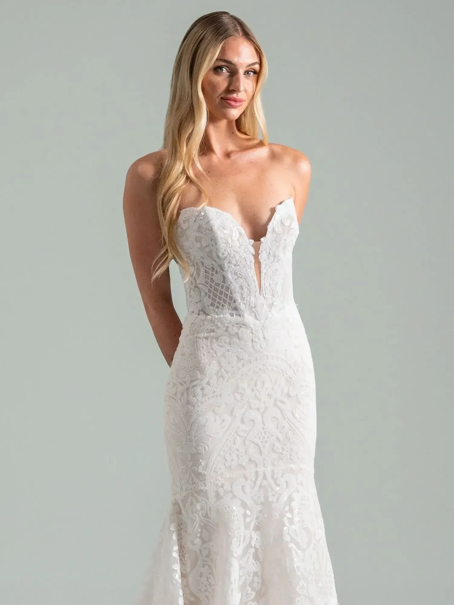A woman wearing a strapless, white wedding gown with intricate lace detailing and a flowing skirt. She has long, straight hair and is standing with her hands behind her back, facing forward.