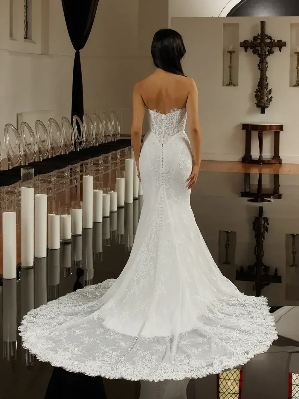 A woman in a white wedding dress with a fitted silhouette and lace details, standing with her back to the camera in a decorated indoor space. The floor is reflective, and there are candles in the background.