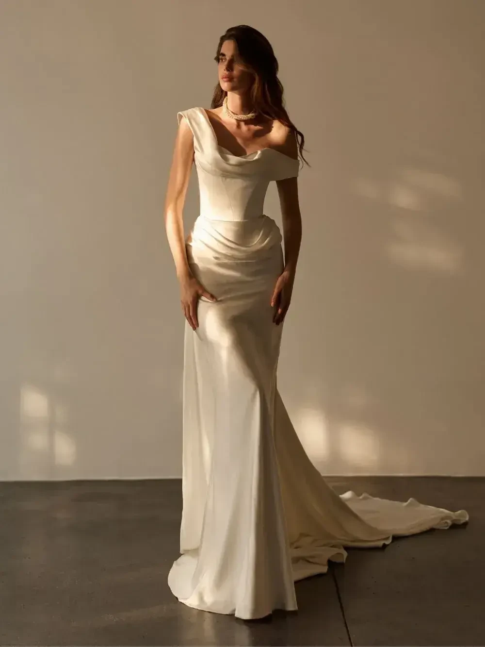 A model wearing a formal white gown with off-the-shoulder sleeves, posed against a light-colored wall.