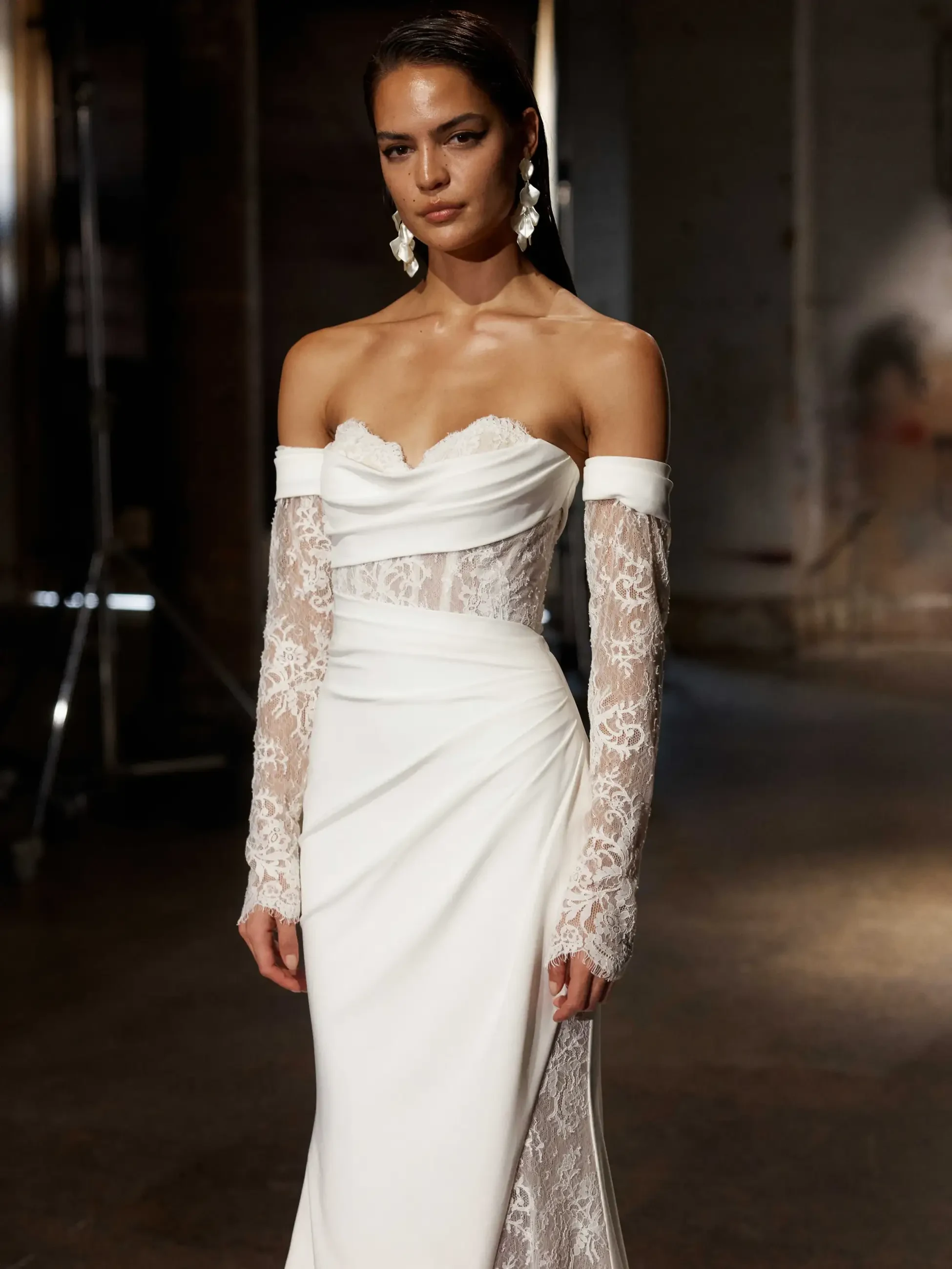 A model wearing a white off-the-shoulder gown with lace sleeves and detailing, standing in an industrial setting.