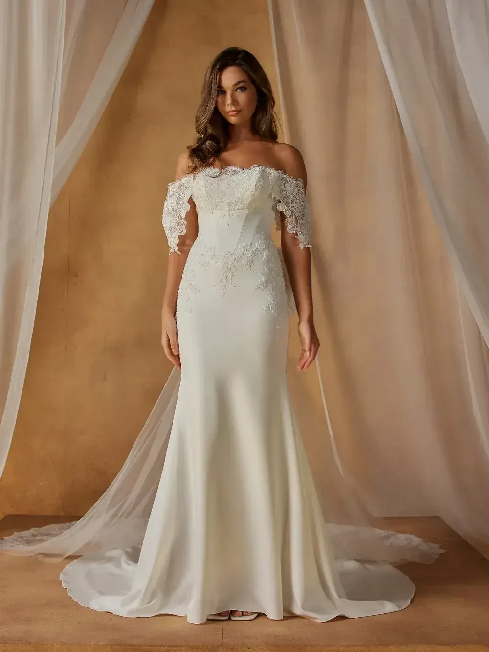 A model in an elegant off-the-shoulder wedding dress with lace sleeves, standing against a soft, neutral backdrop.