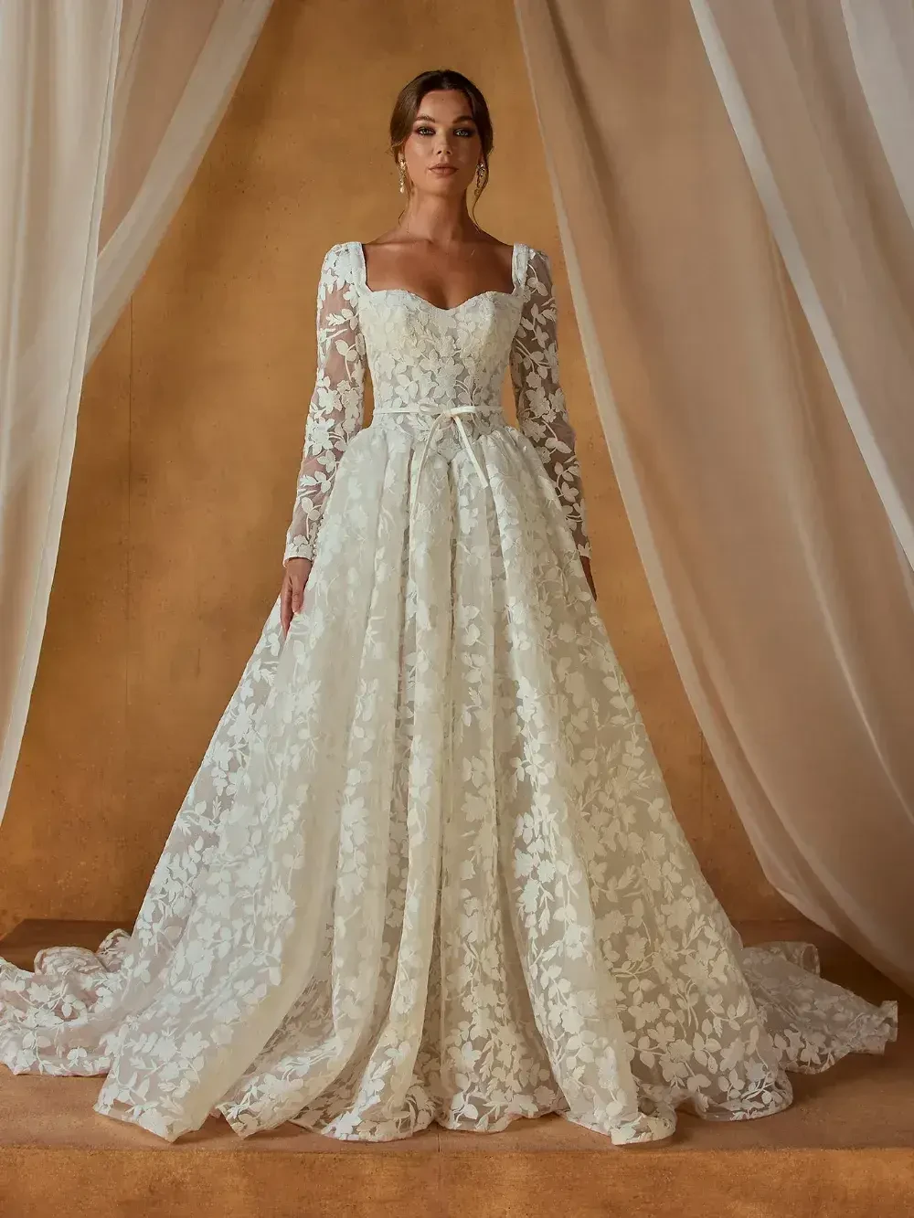 A model wearing a long-sleeved, ivory wedding gown with floral lace details and a full skirt, standing against a neutral background.
