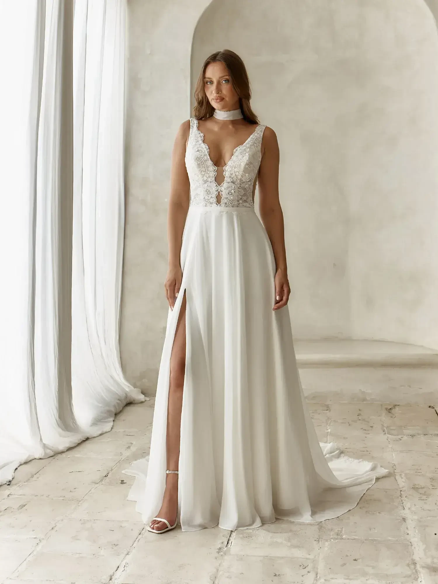 A woman wearing a white wedding dress with a deep V-neck, intricate lace detailing at the top, and a thigh-high slit. She stands in a softly lit room with neutral-colored walls and draped fabric.