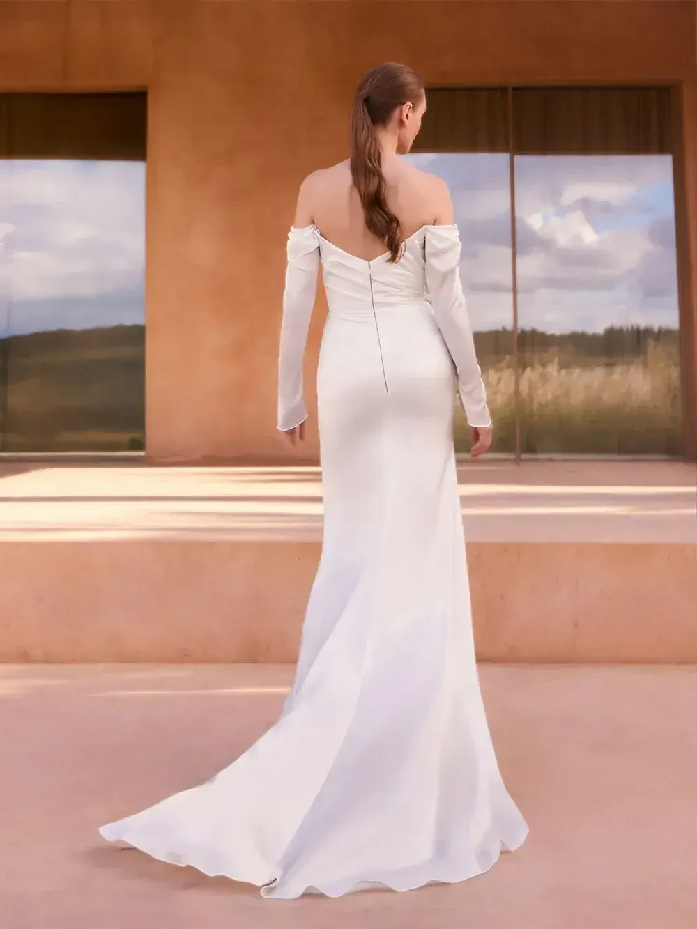 A woman wearing a long, off-the-shoulder white dress, standing with her back to the camera, with a backdrop of smooth surfaces and reflections.